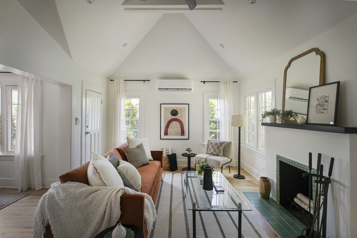 Bright, modern living room with vaulted ceilings, hardwood floors, a rust-colored sofa, fireplace, large windows, and white double entry doors.