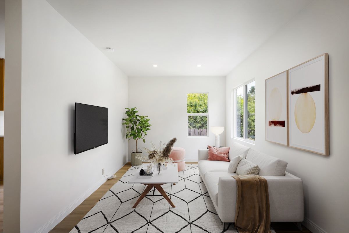 Staged contemporary living room featuring a white sectional sofa, wall-mounted television, geometric rug, modern artwork, coffee table, and large windows with abundant natural light.
