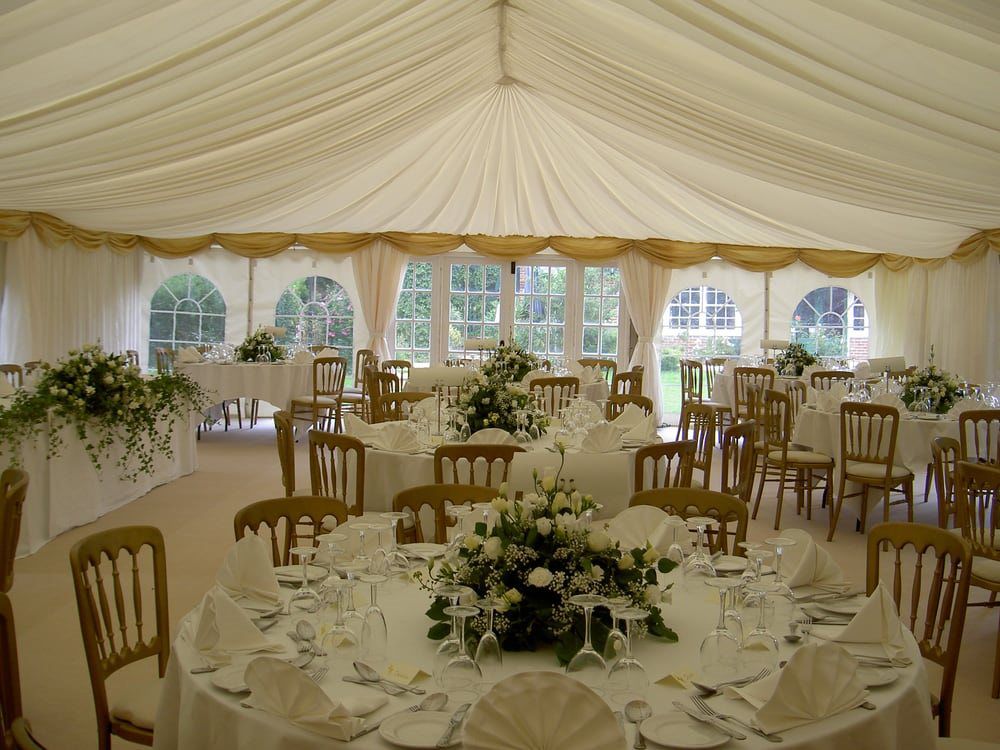 Elegant wedding reception setup inside a marquee with white drapes, round tables, wooden chairs, and floral centerpieces for your wedding inspiration.