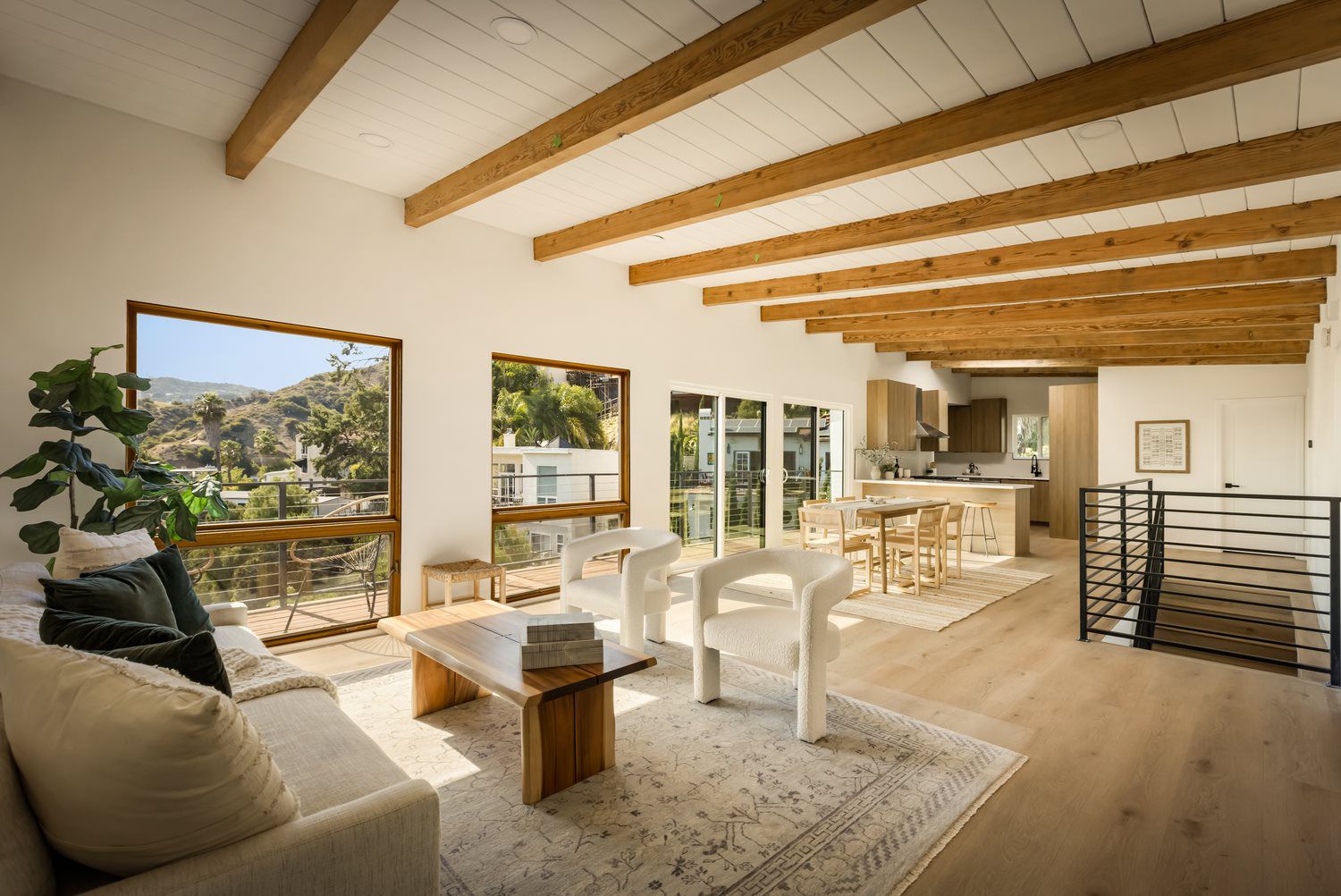 Bright modern living room with exposed wood beams, large windows, and an open kitchen dining area.