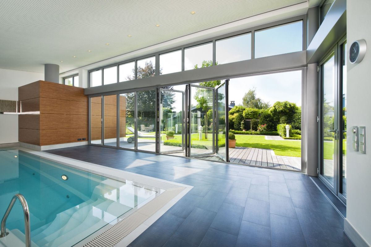 All Weather Series 9200 aluminum bi-fold glass doors open an indoor swimming pool area to an outdoor deck and landscaped garden.