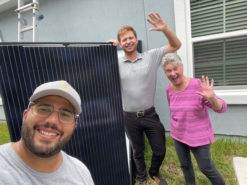 Three happy people stand outside a home. One smiles, holding the camera, while two others wave while holding a solar panel in the back.