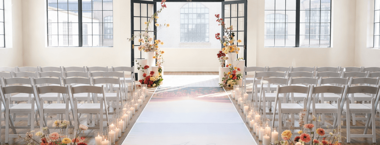 Modern indoor wedding ceremony setup with white chairs, candle-lined aisle, floral arrangements, and black-framed arched windows in a bright loft-style venue.