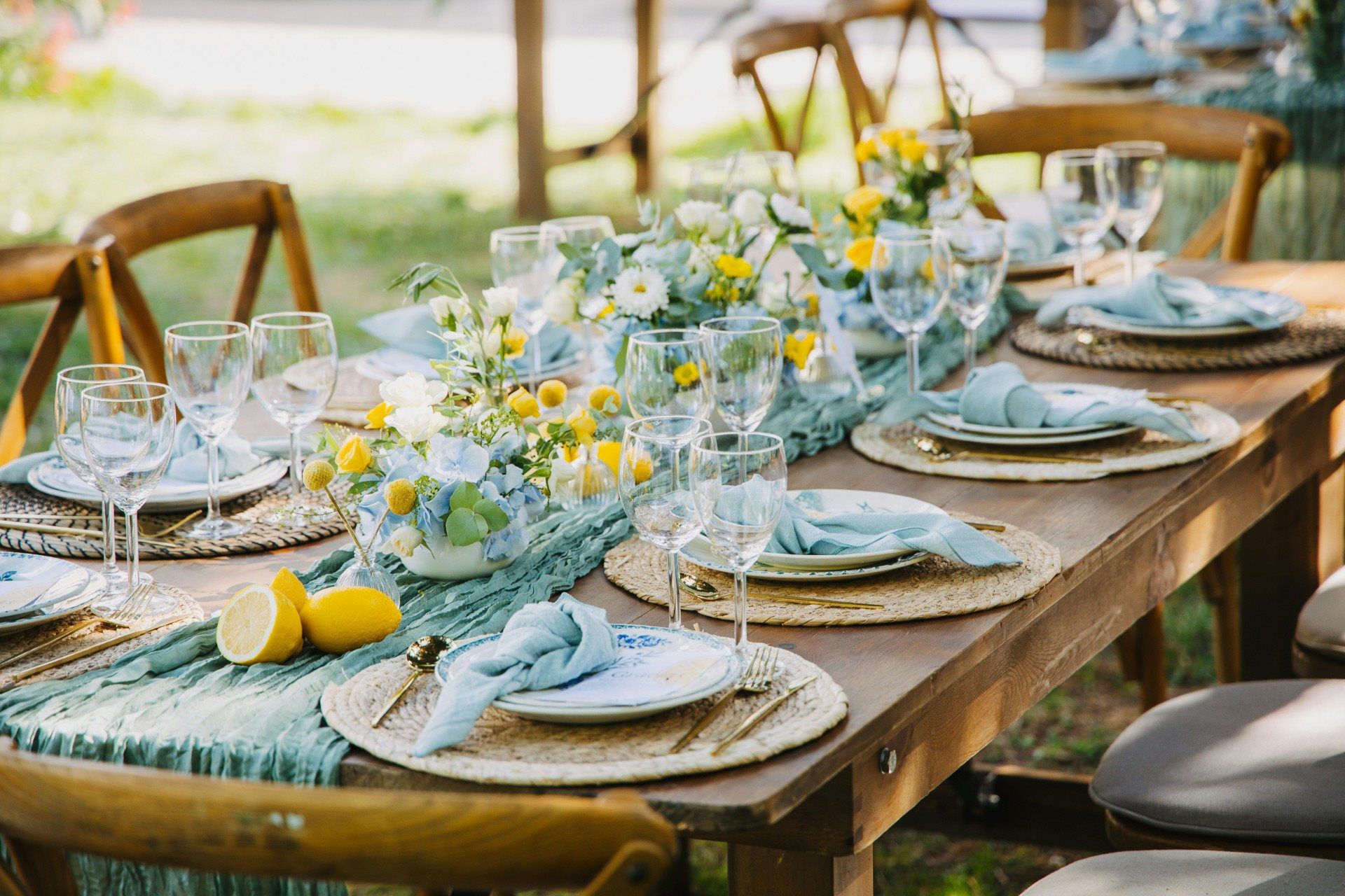 A high-quality, close-up view of an outdoor wedding table setting featuring a specific Amalfi-inspired theme. The wooden harvest table is decorated with a sage green textured gauze runner and vibrant yellow lemons scattered throughout. A white table number card sits in a gold stand, featuring blue "Table 2" script and lemon illustrations. The place setting includes a woven straw charger, a white ceramic plate with blue floral patterns, a light blue knotted linen napkin, and gold flatware. Small floral arrangements of light blue hydrangeas and yellow roses add a sense of calm confidence and exclusive, artisanal detail to the scene.