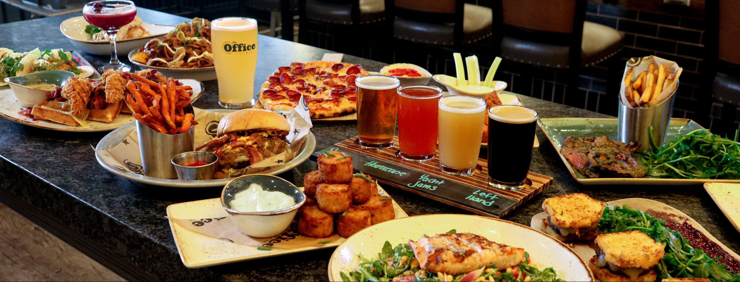 picture of multiple food and drink items at The Office including beer, a cocktail, pepperoni pizza, wings, sweet potato nachos, Tavern burger and fries, jammin’ sliders, tater tots and more.