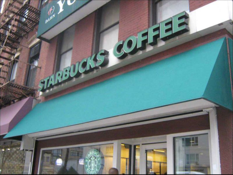 A bustling Starbucks location featuring a stylish green awning with the Starbucks logo displayed prominently. This commercial awning provides shade for the storefront’s entrance and creates a welcoming ambiance for customers. The awning is sturdy, de