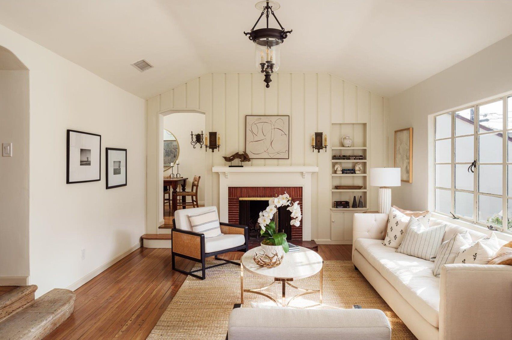 Bright living room with white sofa, wood floors, fireplace, built-in shelves, and large window letting in natural light.