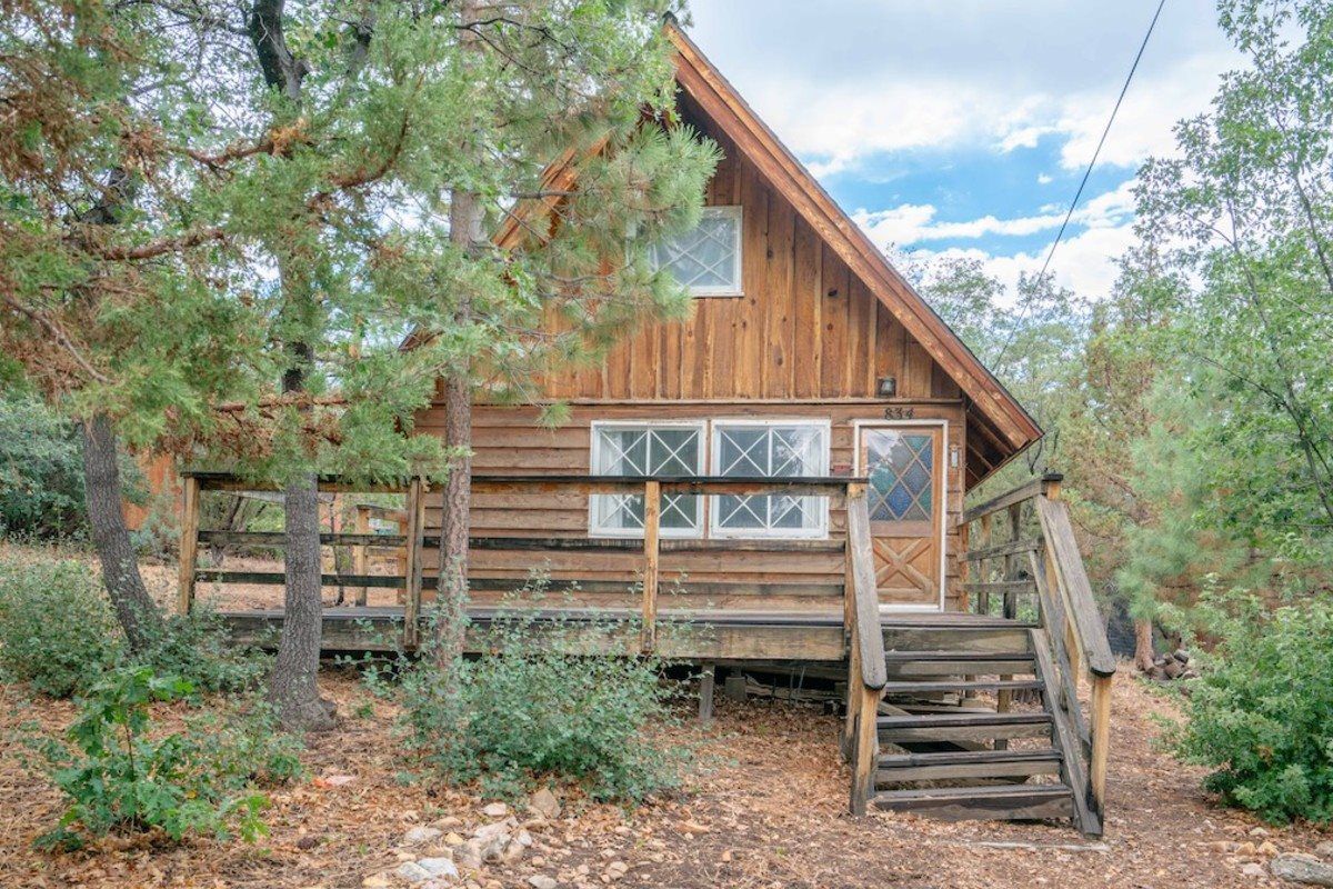 Rustic A-frame wooden cabin with a front porch and steps, surrounded by mature trees and dense greenery.
