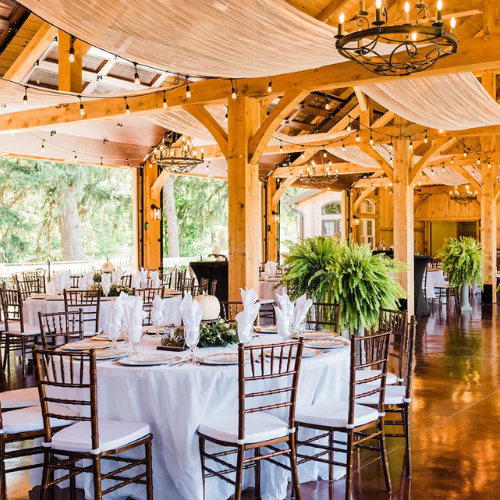 A decorated event space by Weddings & Events by K'Mich, featuring wooden beams, round tables with white cloths and napkins, elegant chairs, chandeliers, and potted ferns. Large windows reveal greenery outside—a perfect Philadelphia setting curated by an expert wedding planner at Weddings & Events by K'Mich